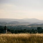 Tranquil landscape of Kamienna Góra, Poland, featuring lush fields, hills, and a distant church under a sunset sky.