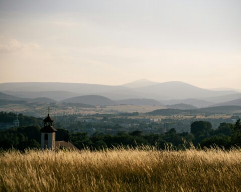 Tranquil landscape of Kamienna Góra, Poland, featuring lush fields, hills, and a distant church under a sunset sky.
