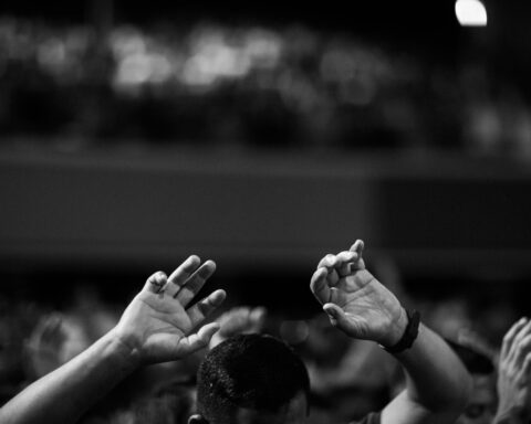 A black and white photo of hands raised in a crowd during a concert or religious gathering indoors.