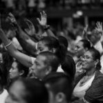 Black and white photo of a diverse crowd with raised hands at an indoor event.