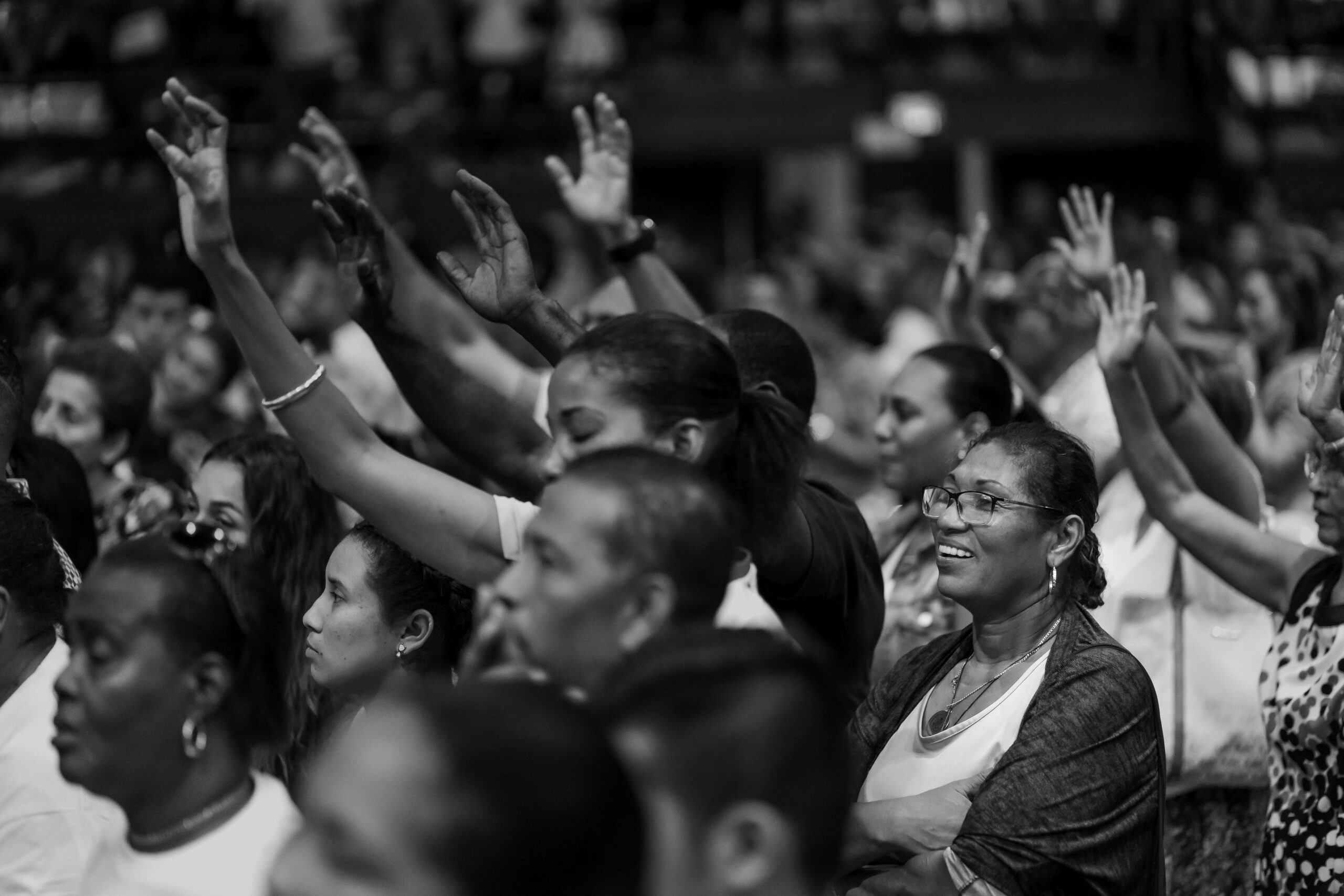 Black and white photo of a diverse crowd with raised hands at an indoor event.
